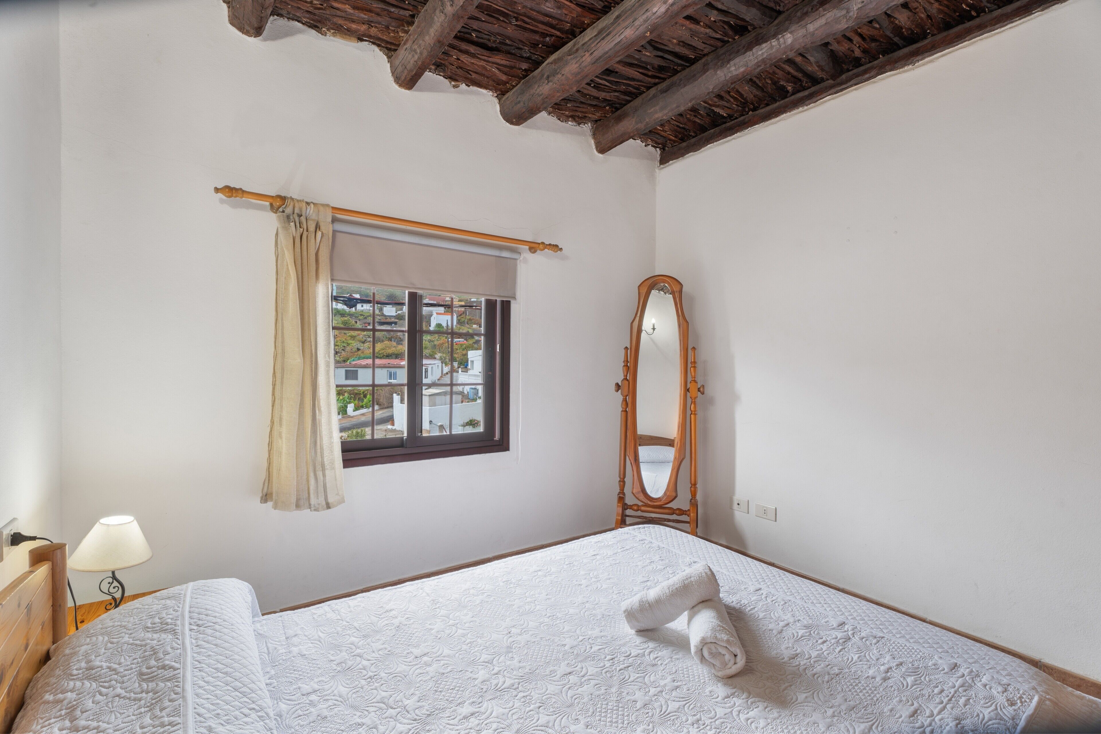 A bedroom with a bed, a window with curtains, a wooden ceiling, and a mirror.