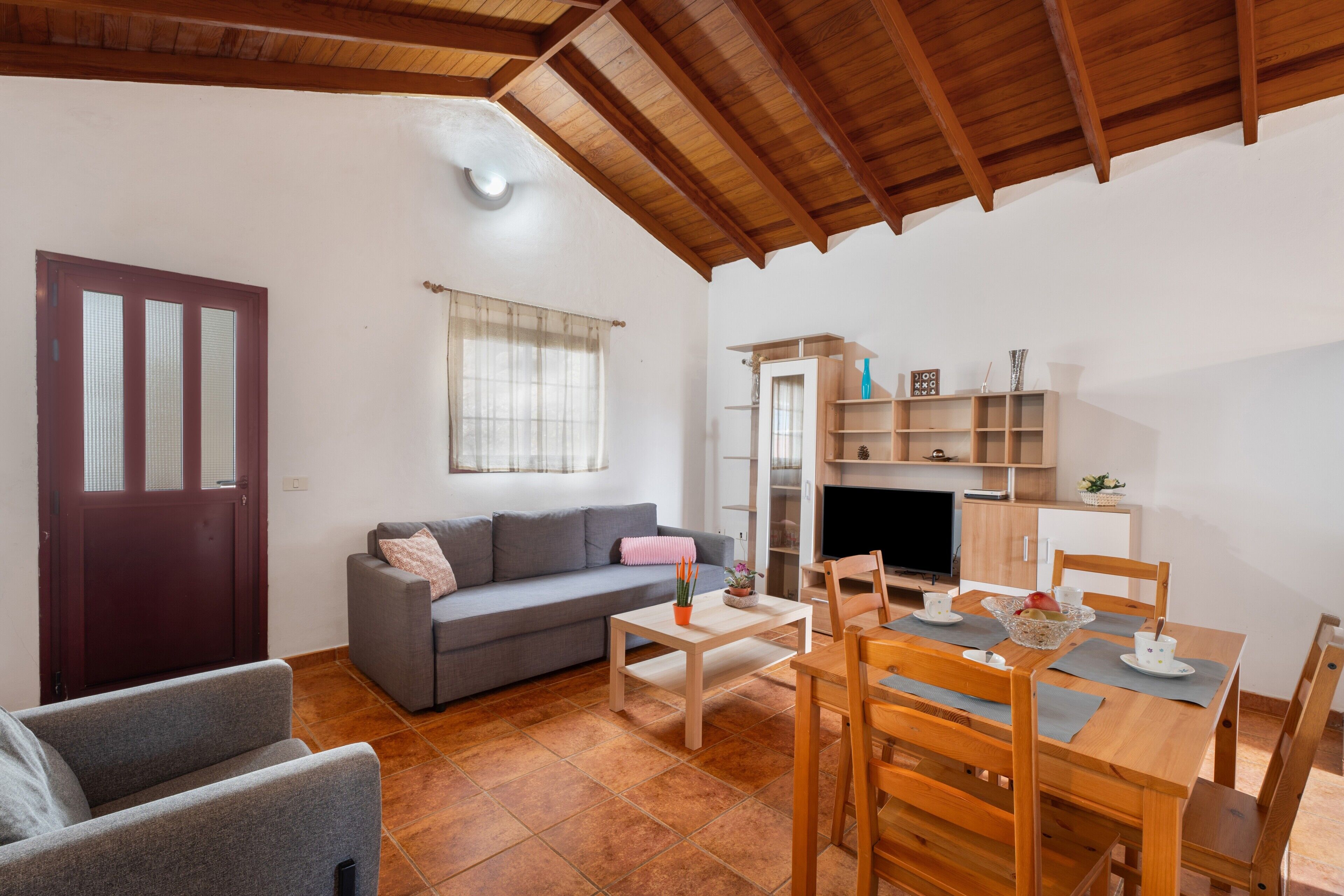 A living room with a sofa, a dining table with chairs, a TV, and a kitchen area with shelves and a cabinet.