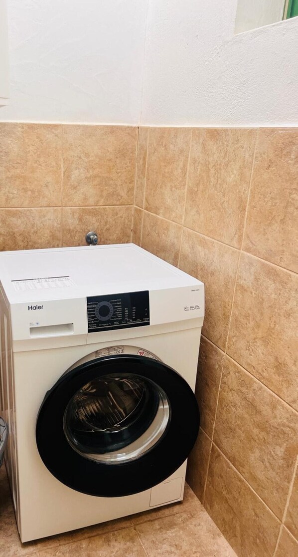 En lille, hvid Haier vaskemaskine står i et hjørne med beige flisegulve.