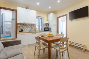 A compact kitchen with white cabinets, a dining table with chairs, a TV mounted on the wall, and a view of the bathroom through an open door.