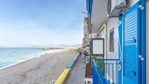 Un hotel frente al mar con balcón y vistas a la playa, una puerta azul y montañas a lo lejos.