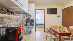 Cocina pequeña con tostadora, cafetera y un electrodoméstico rojo, que da paso a un dormitorio con televisor y puerta de madera.