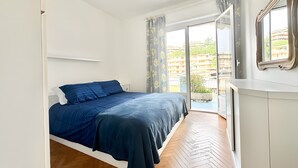A bedroom with a bed, a ceiling fan, a window with curtains, and a view of buildings outside. - Apartment 'Flaminia' with Balcony and Wi-Fi (Sanremo)