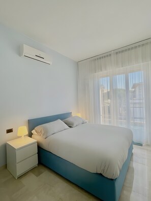 A bedroom with a bed, nightstand, lamp, air conditioning unit, and a window with sheer curtains.