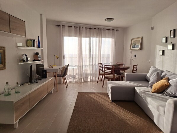 Una sala de estar moderna con un sofá, una mesa de comedor y sillas. Hay un televisor sobre un mueble, una estantería y una ventana con cortinas.