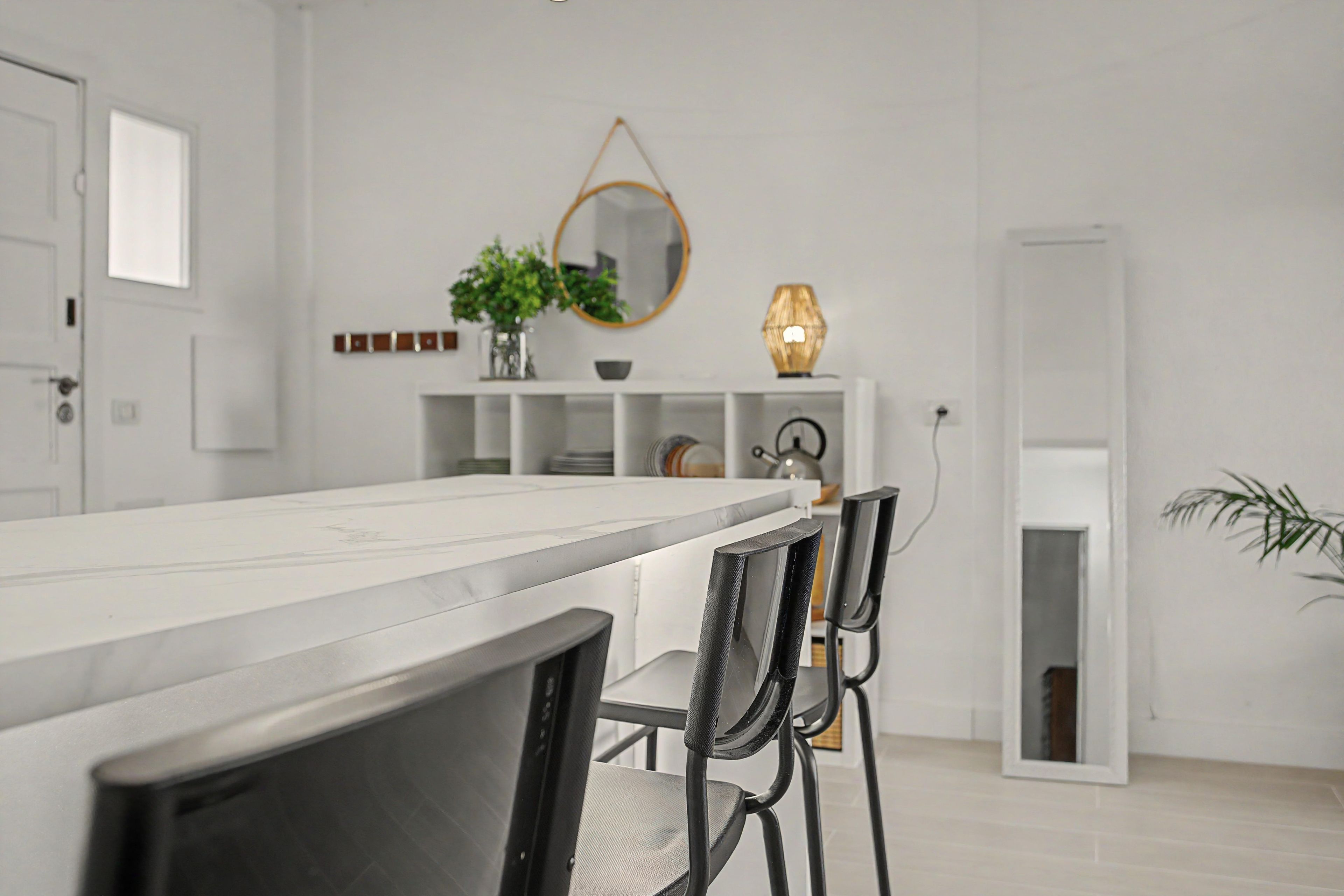 Un espace repas moderne avec une table blanche, des chaises noires, un miroir, une plante et une lampe.