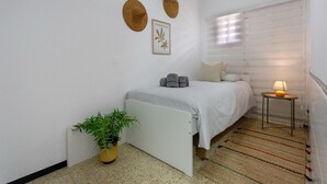 Un dormitorio con una cama, una mesita de noche con lámpara, una planta en maceta y dos sombreros colgados en la pared.