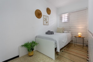 Un dormitorio con una cama, una mesita de noche con lámpara, una planta en maceta y dos sombreros colgados en la pared.