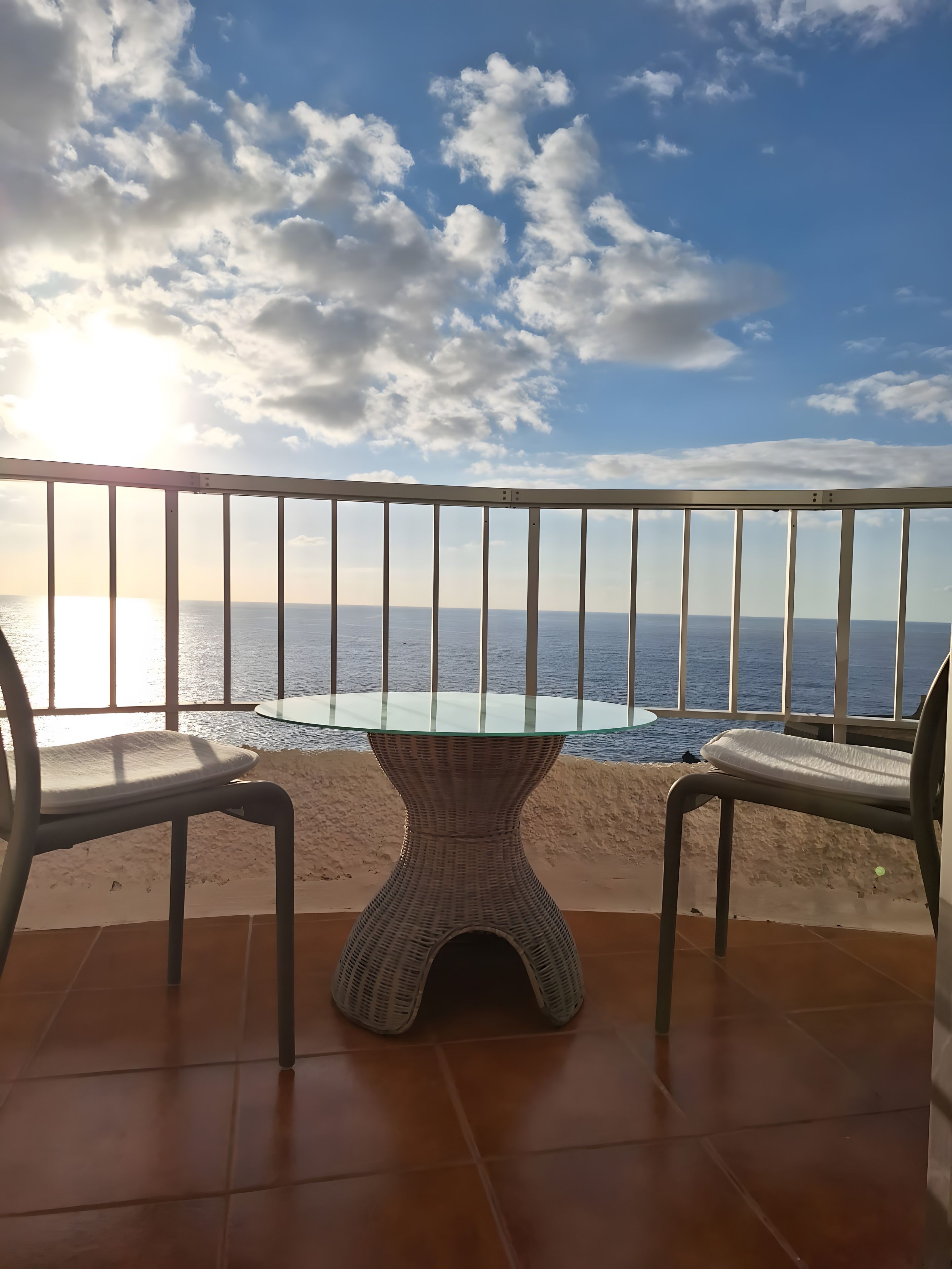 Un balcon doté d’une table à plateau en verre et de deux chaises, offrant une vue sur une plage et la mer.