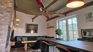 Een rustiek interieur met zichtbare houten balken, een bakstenen muur, een houten tafel, een zwarte bank en een groot raam.