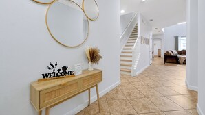 A hallway with a wooden console table, a welcome sign, and three wall-mounted mirrors. A staircase is visible in the background leading to another room.