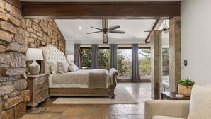 A bedroom with a stone wall, a bed, a nightstand, a ceiling fan, and a view through large windows. - Grotto @ Southern Hills W/Pool & Hot Tub (Tulsa)