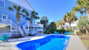 A swimming pool with a curved edge, surrounded by a deck with chairs and tables. There are palm trees and a building with a balcony in the background. - Spacious 8BR/6BA Duplex with Private Pool – Walk to the Beach – Sleeps 22 (North Myrtle Beach)