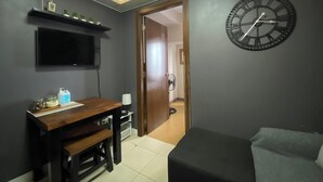 Una habitació amb escriptori de fusta, rellotge negre a la paret, televisió penjada sobre l'escriptori i porta que dona a una altra habitació.