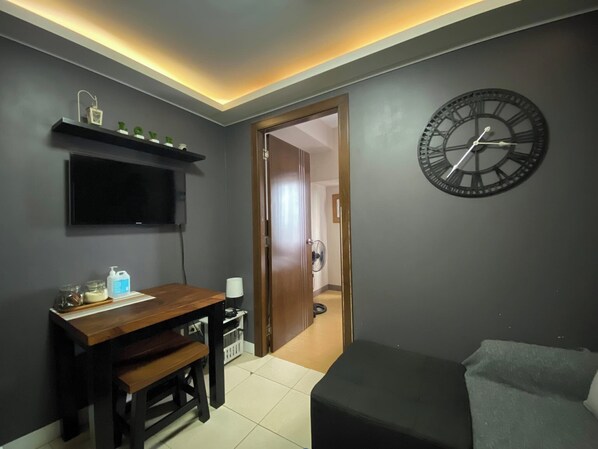 Une pièce avec un bureau en bois, une horloge noire accrochée au mur, un téléviseur fixé au-dessus du bureau et une porte donnant sur une autre pièce.