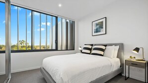 A modern bedroom with a large window, a bed with white bedding, a bedside table with a lamp, and a framed picture on the wall.