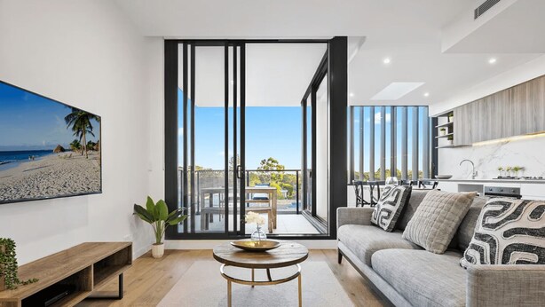 A modern living room with a large sliding glass door leading to a balcony, a grey sofa, a wooden coffee table, and a TV mounted on the wall.