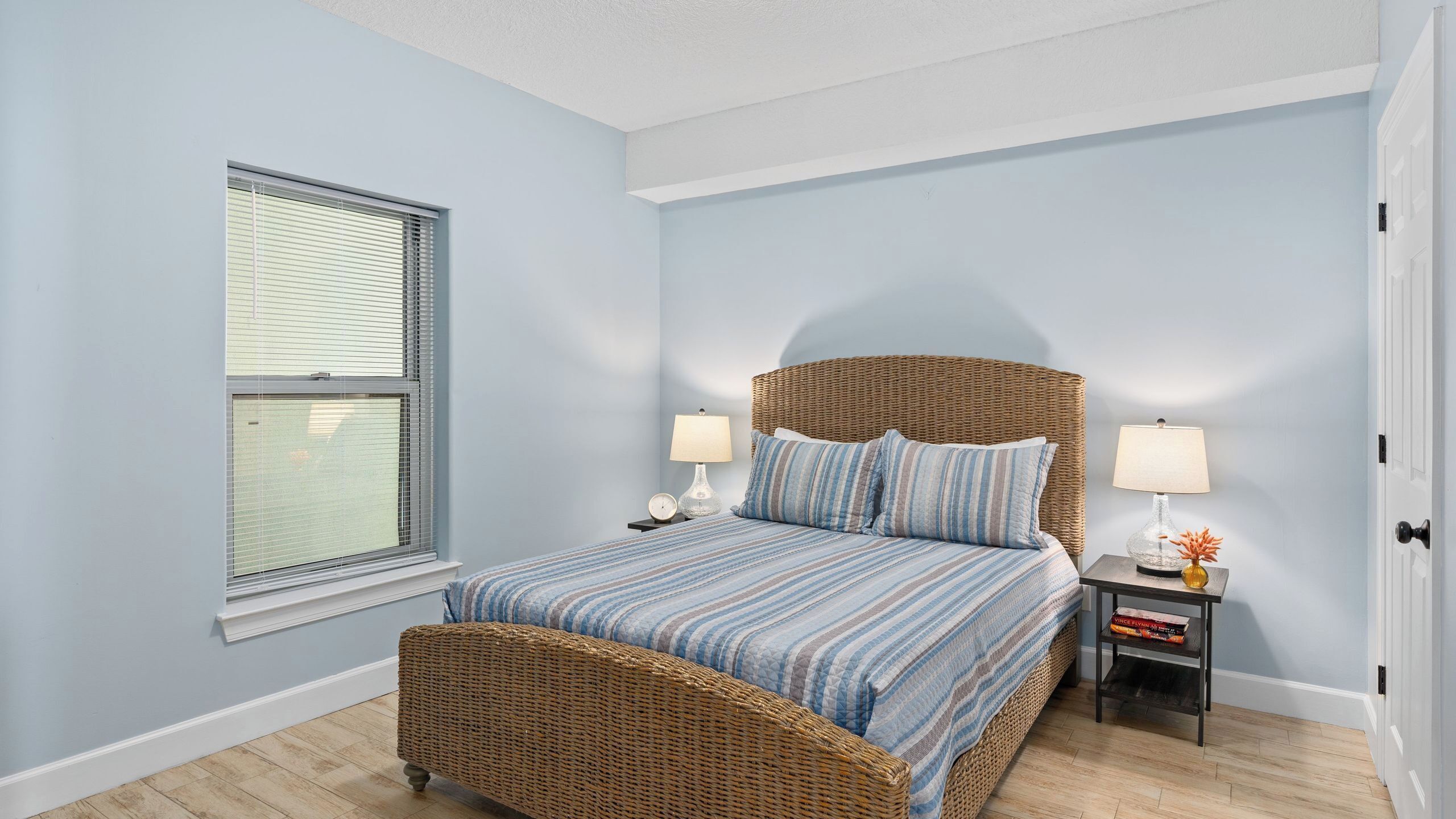 A bedroom with a wicker bed, two bedside tables with lamps, a window with blinds, and a door.