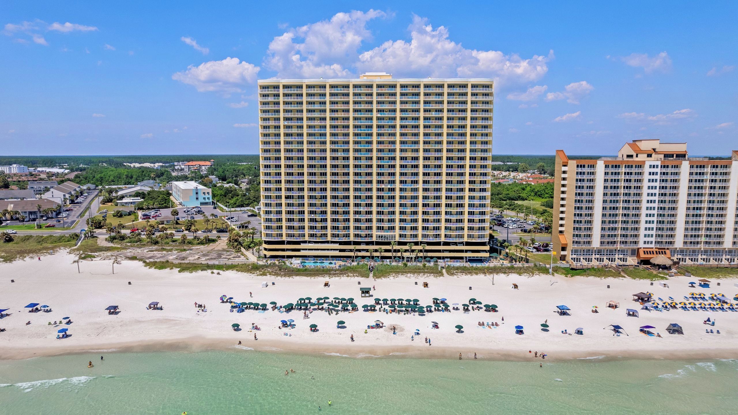 Un hotel grande con varios pisos y balcones, una playa con numerosos sombrillas y sillas de tomar el sol, y una vista despejada del océano.