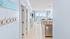 A modern kitchen with a view to the outside, a dining area with a blue couch, and a hallway with a sign reading "PANAMA CITY BEACH". - Beachfront Condo w/ Ocean Views + Pool Access + Beach Gear Emerald Isle 808 by Avantstay (Panama City Beach)
