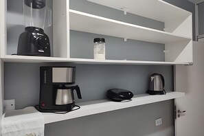 Una kitchenette con cafetera, hervidor y una botella de agua sobre un estante blanco.
