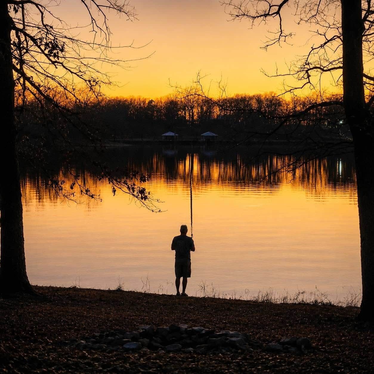 A person fishing by a calm lake at sunset with trees and a distant building.
