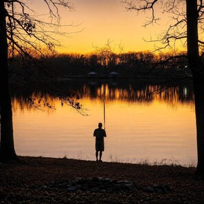 Une personne est en train de pêcher au bord d’un lac paisible, au coucher du soleil, avec des arbres et un bâtiment visible au loin.