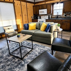 A cozy living room with a sofa, coffee table, and ottoman, a kitchen with wooden cabinets and appliances, and a window with blinds. - Playin Hooky on Lake Fork-Waterfront-Boat Ramp (Quitman)