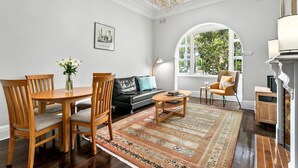 A living room with a wooden dining table, chairs, a black sofa, a patterned rug, and a large arched window with a view of greenery.