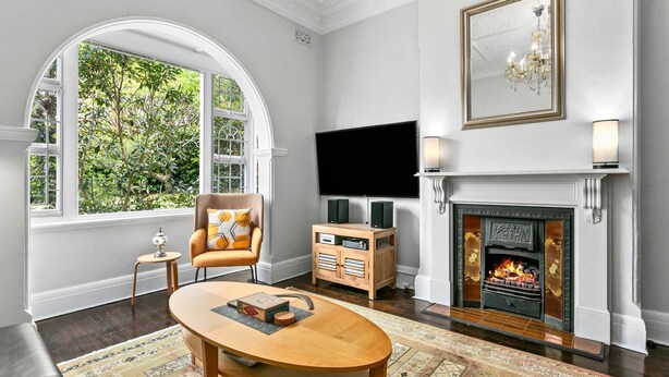A living room with a fireplace, a television, a comfortable armchair, and a large window with a view of greenery.