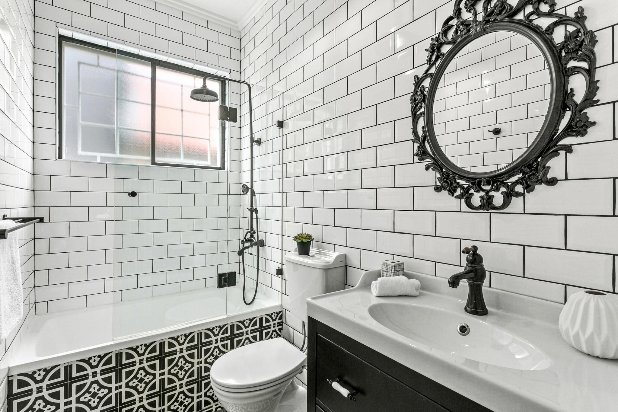 A bathroom with white subway tiles, a black and white patterned tile accent, a round mirror with an ornate frame, a white sink, and a toilet.