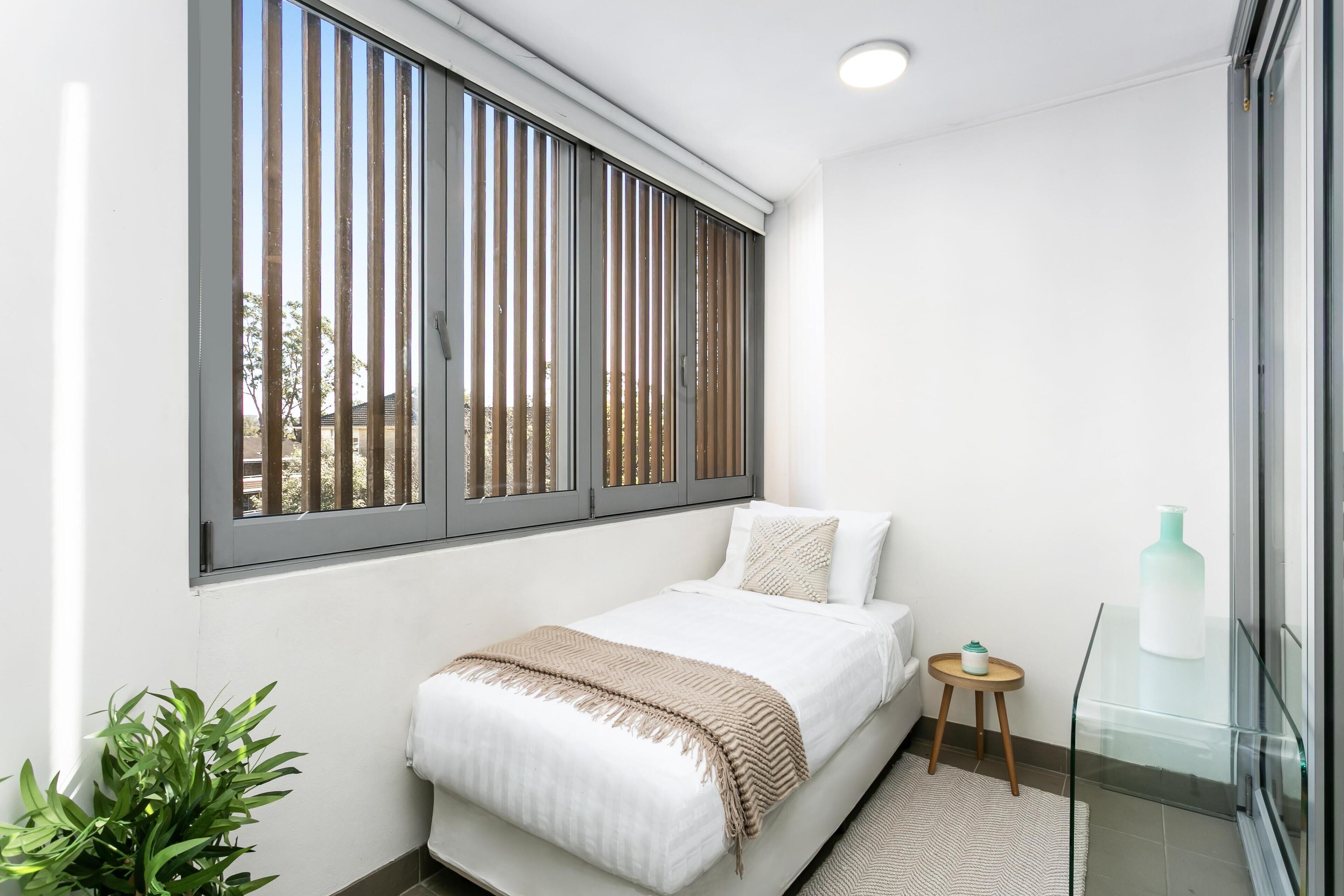 Un dormitorio con cama, una mesita de noche pequeña, una planta en maceta, una ventana con persianas y una mesa de cristal.