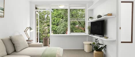 Ein modernes Wohnzimmer mit einer Couch, einem Fernseher auf einem Regal, einem Fenster mit Blick auf Grünflächen und einer Tür.