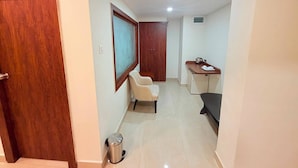 A hallway with a wooden door, a framed mirror, a small seating area, a kitchenette, and a trash bin.