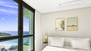 A hotel room with a large window offering a view of the ocean and a balcony. There are two framed pictures on the wall and a bedside table with a lamp. - A1302 (Cupecoy)