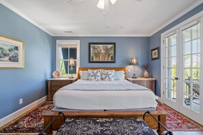 A bedroom with a large bed, wooden headboard, two bedside tables with lamps, a window with blinds, and a door leading outside.