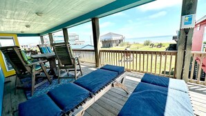 A covered outdoor area with wooden lounge chairs and a table, overlooking a beach and ocean. - Beachfront 3BR/2BA – Steps to the Sand, Huge Deck (Bolivar Peninsula)