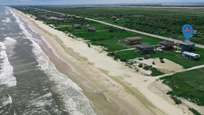An aerial view of a coastal area with a beach, green fields, and several buildings. - Beachfront 3BR/2BA – Steps to the Sand, Huge Deck (Bolivar Peninsula)