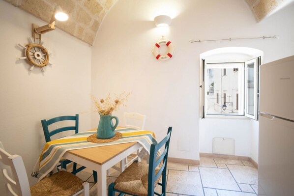 A dining area with a table, chairs, a vase with dried flowers, a life preserver decoration, and a view through an open door.
