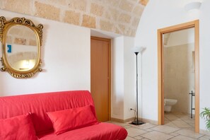 Ein Zimmer mit einem roten Sofa, einem goldfarbenen Spiegel, einer Holztür, einer Stehlampe und einem durchgehenden Tür sichtbar liegenden Badezimmer.
