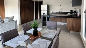 A modern living room with a glass dining table, grey sofas, and a kitchen area with a refrigerator, stove, and sink. - Araquuna gated community, 2 bd condo close to PV, Sayulita, Bucerias (Mezcales)