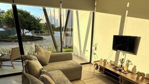 A modern living room with a sectional sofa, a flat-screen TV, and a large window offering a view of the outdoors. - Araquuna gated community, 2 bd condo close to PV, Sayulita, Bucerias (Mezcales)