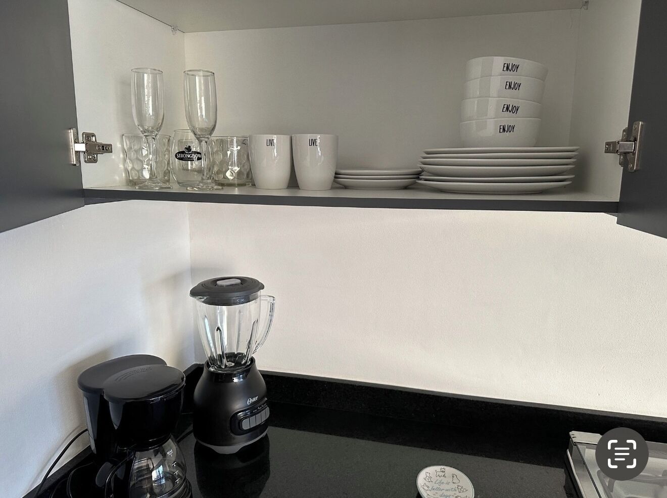 A kitchen with a black countertop, a blender, a water bottle, and a coffee maker. There are also wine glasses, cups, and plates on a shelf above the counter.