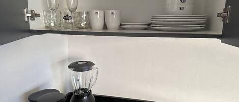 A kitchen with a black countertop, a blender, a water bottle, and a coffee maker. There are also wine glasses, cups, and plates on a shelf above the counter.