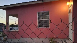 Un edificio rosa con una ventana protegida por barrotes y una puerta, rodeado por una cerca de malla metálica.