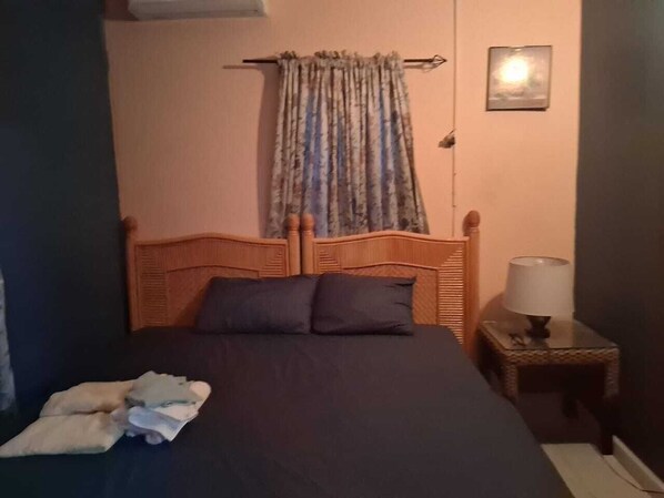 A bedroom with a wooden headboard, a bed with dark bedding, a nightstand with a lamp, and a window with a curtain.