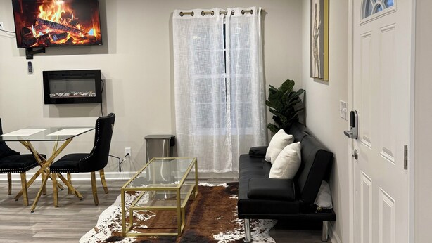 A modern living room with a flat-screen TV, ceiling fan, dining area with a glass table and chairs, a black sofa, and a cowhide rug.