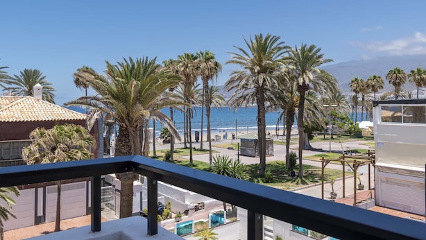 A balcony with a view of a beach, palm trees, and a clear sky. - Parque Santiago II - 2 Bed Sea View (Playa de Las Americas)