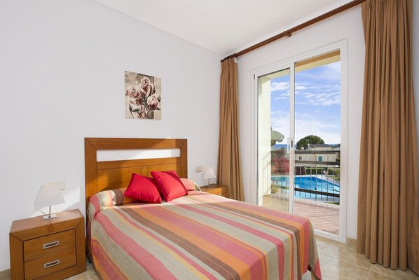 Habitación de hotel con una cama grande, una mesita de noche, una lámpara, un cuadro y un ventanal corredero a una zona de piscina.