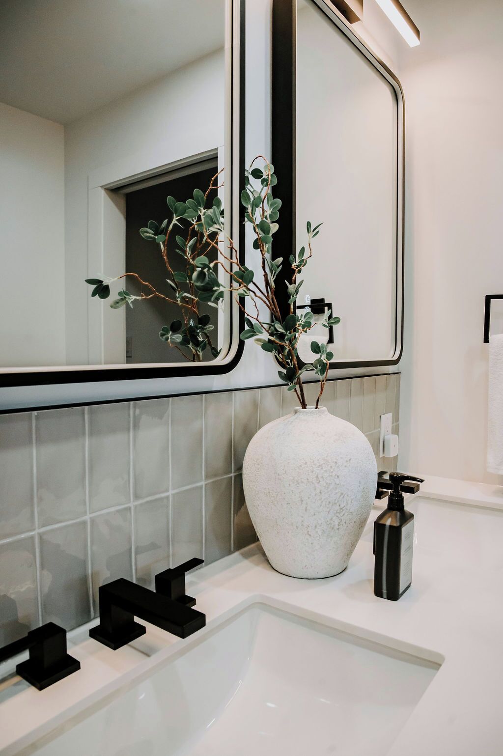 A modern bathroom with a white sink, black faucets, a large mirror, and a white vase with green foliage.